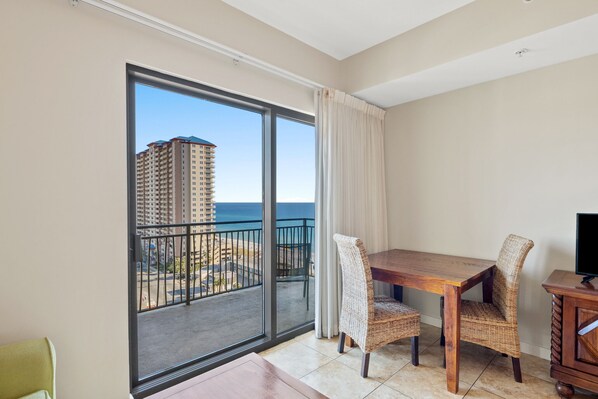 A balcony with a view of a high-rise building and the ocean. There is a wooden table with two wicker chairs and a TV on a wooden cabinet.