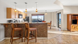 A modern kitchen with wooden cabinets, a central island with bar stools, pendant lighting, and a view to the outside. - Origin 1235 - 2 Bedroom - Stunning Gulf Views (Panama City Beach)