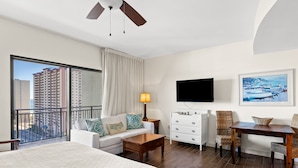 A hotel room with a large bed, a sofa, a desk, and a TV. There is a balcony with a view of tall buildings. - Origin 1329 - Studio King Gulf View - Sunrise Views (Panama City Beach)