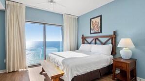 Un dormitorio con una cama grande, una mesita de noche con lámpara, un banco y vista al océano a través de un ventanal.