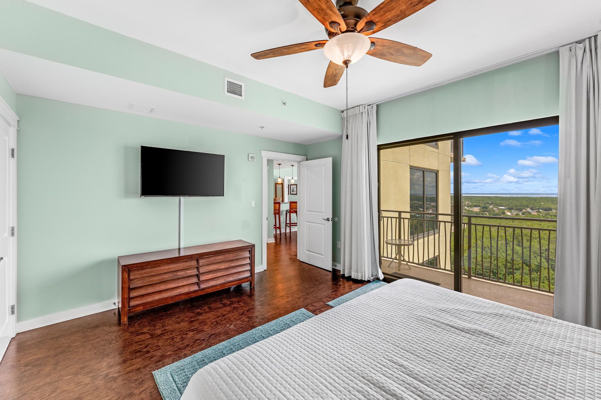 A bedroom with a large bed, a ceiling fan, a TV, and a balcony with a view.