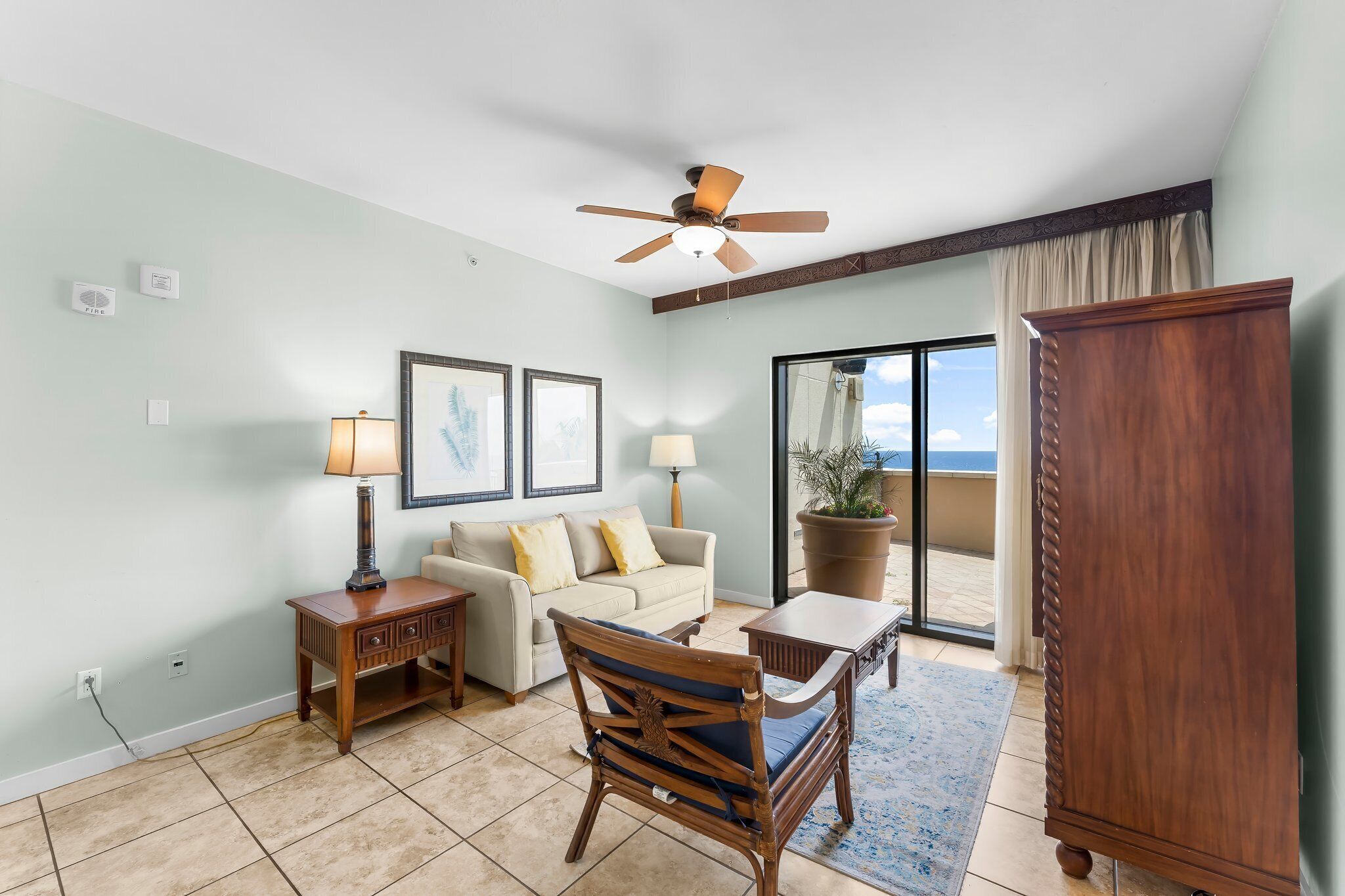 A living room with a sofa, a wooden chair, a coffee table, a ceiling fan, and a view through a glass door to the outside.