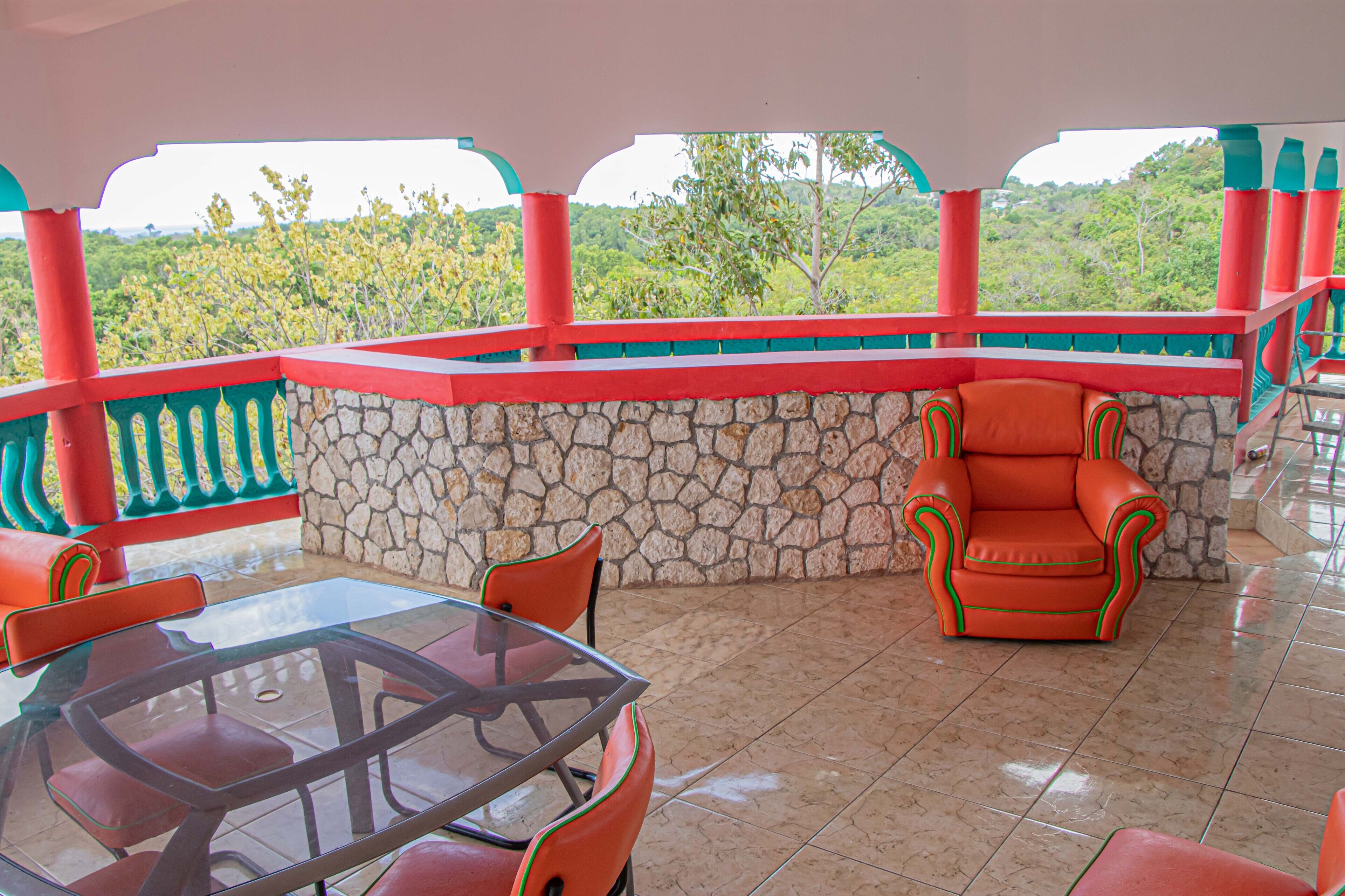 Un patio cubierto con una mesa de vidrio y sillas rojas, paredes de piedra y vista a zonas verdes.