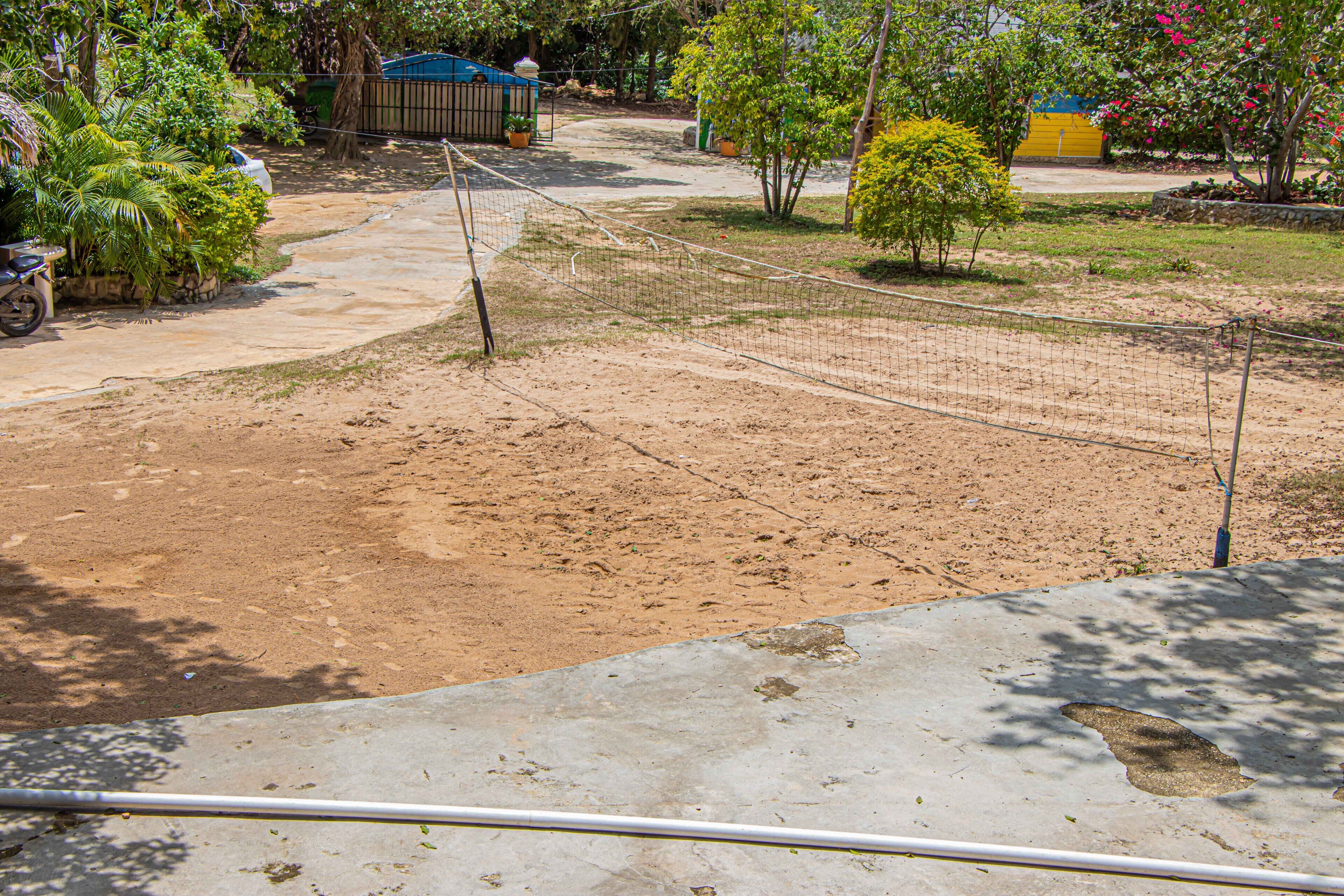 A sandy outdoor area with a volleyball net, surrounded by greenery and a few potted plants.