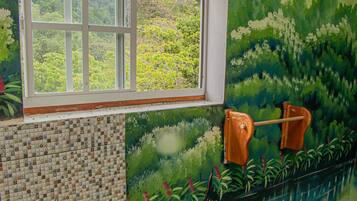 Bathroom features a unique artistic design with a large window offering a view of lush greenery outside, mosaic tile walls, and a vibrant mural.