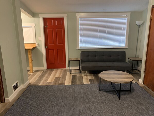A living room with a red door, a grey sofa, a round coffee table, and a window with blinds.