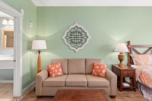 Une pièce avec un canapé beige, deux coussins orange, une table de chevet en bois avec une lampe, et un lit avec une literie à motifs.