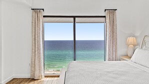 Een slaapkamer met een groot bed, een nachtkastje met een lamp en uitzicht op de oceaan door een schuifdeur.