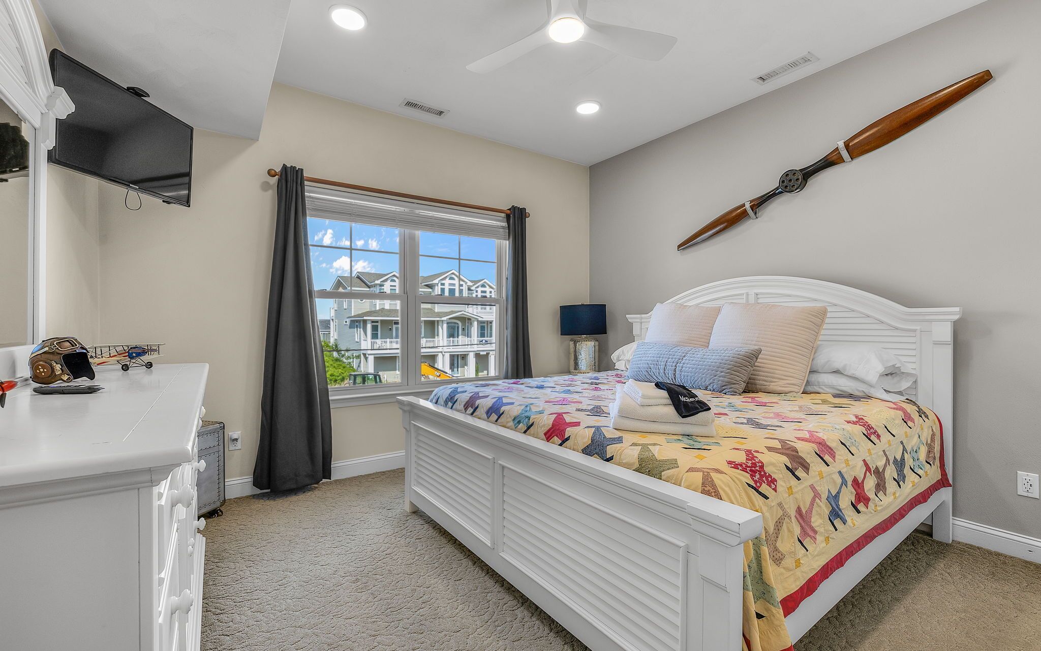 Une chambre à coucher comprenant un lit, un bureau, une fenêtre avec des rideaux, un ventilateur de plafond et un objet en bois fixé au mur.
