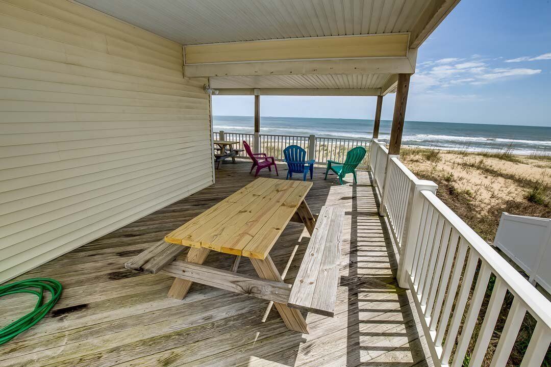 A covered deck with a wooden table and chairs, overlooking a beach.
