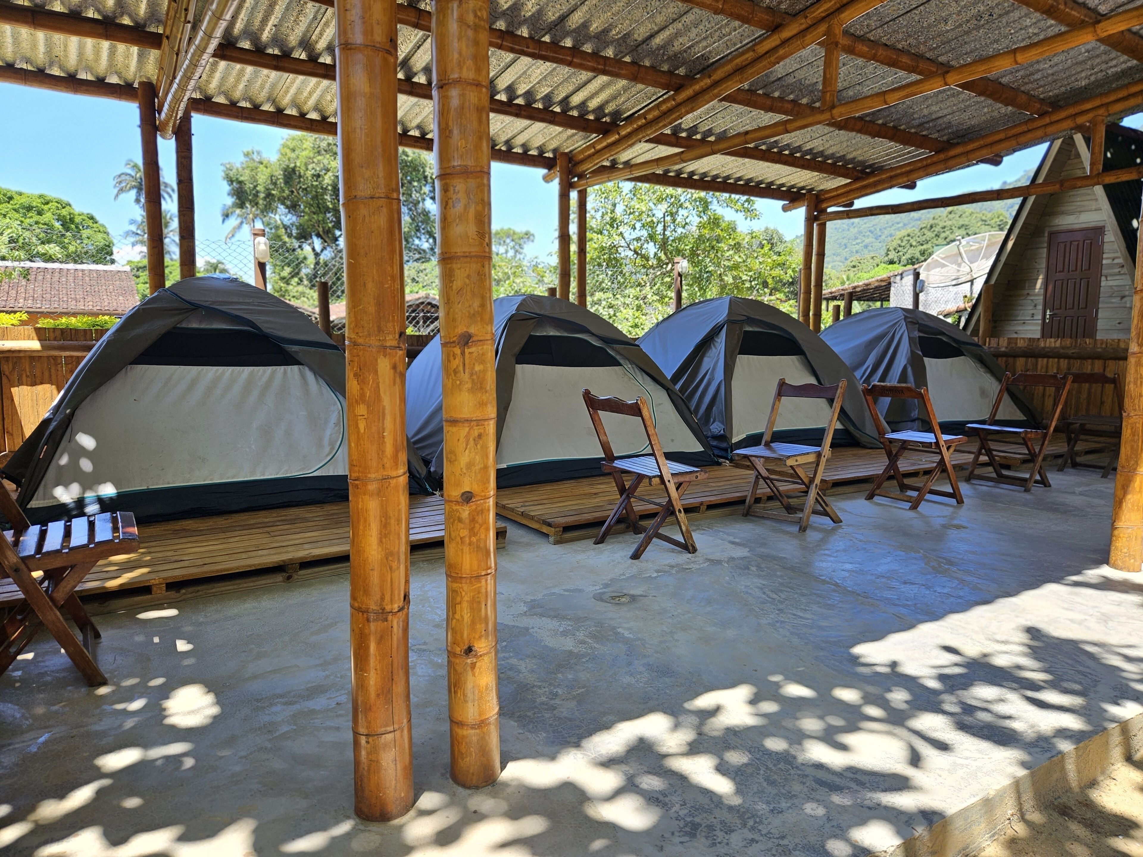 A series of tents with wooden chairs in front, set under a bamboo structure with a view of greenery.