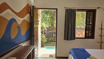 A hotel room with a bed, a TV on a wooden shelf, a door leading to a garden, and a window with curtains.