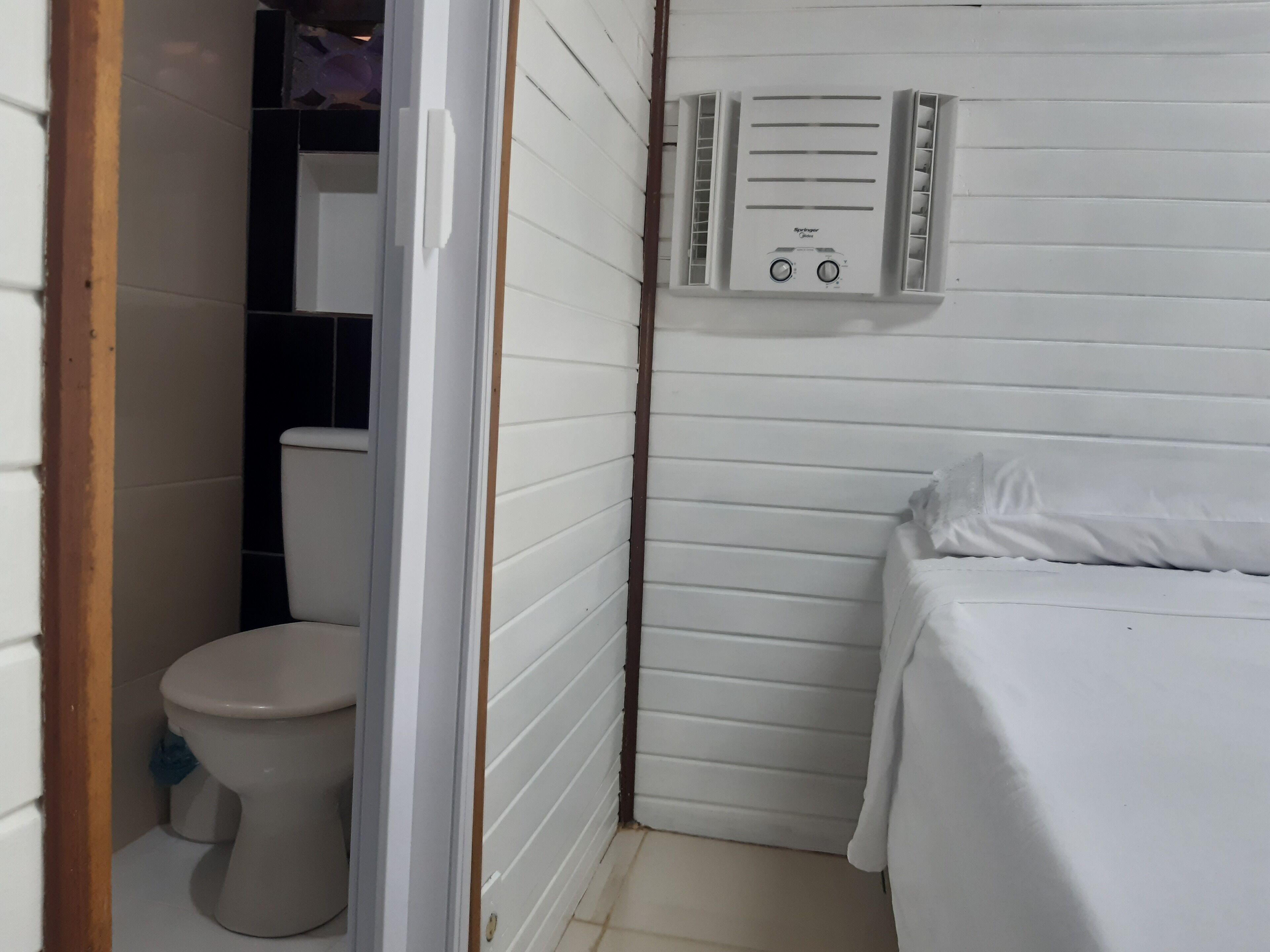 A small, white bathroom with a toilet, a shower area, and a bed.