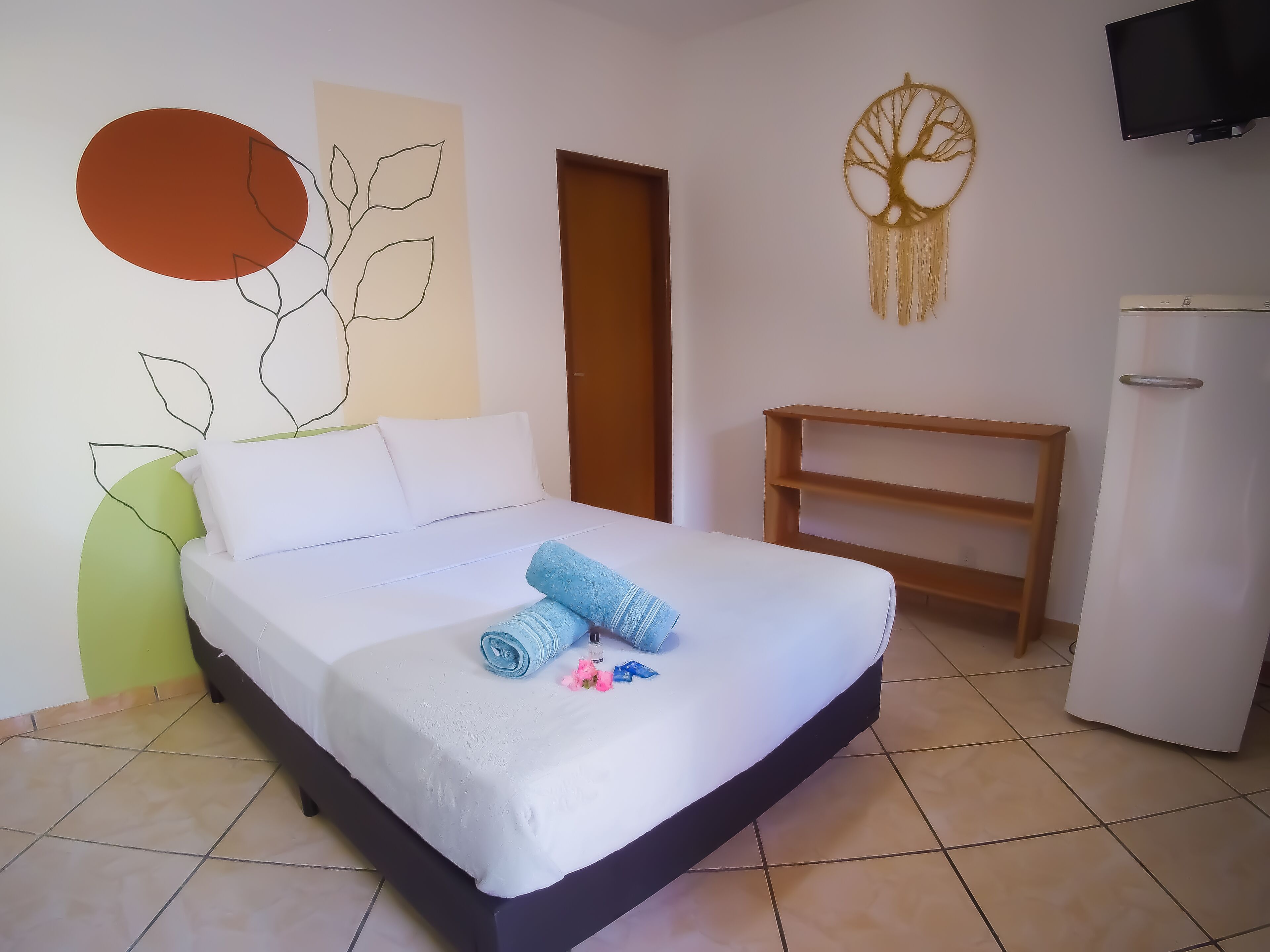 Una cama bien tendida con ropa de cama blanca y azul, una mesita de noche pequeña, un televisor montado en la pared y un adorno en la pared.