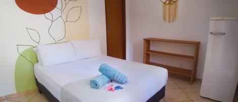 Una cama bien tendida con ropa de cama blanca y azul, una mesita de noche pequeña, un televisor montado en la pared y un adorno en la pared.