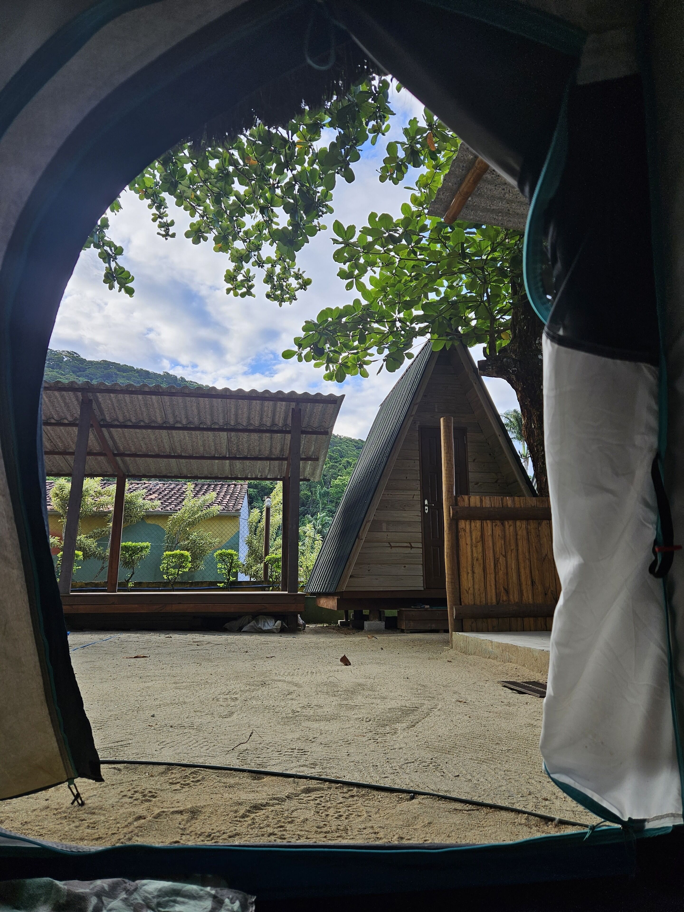A view from inside a tent looking out at a wooden structure with a sloped roof, surrounded by greenery and a clear sky.