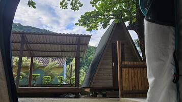 A view from inside a tent looking out at a wooden structure with a sloped roof, surrounded by greenery and a clear sky.