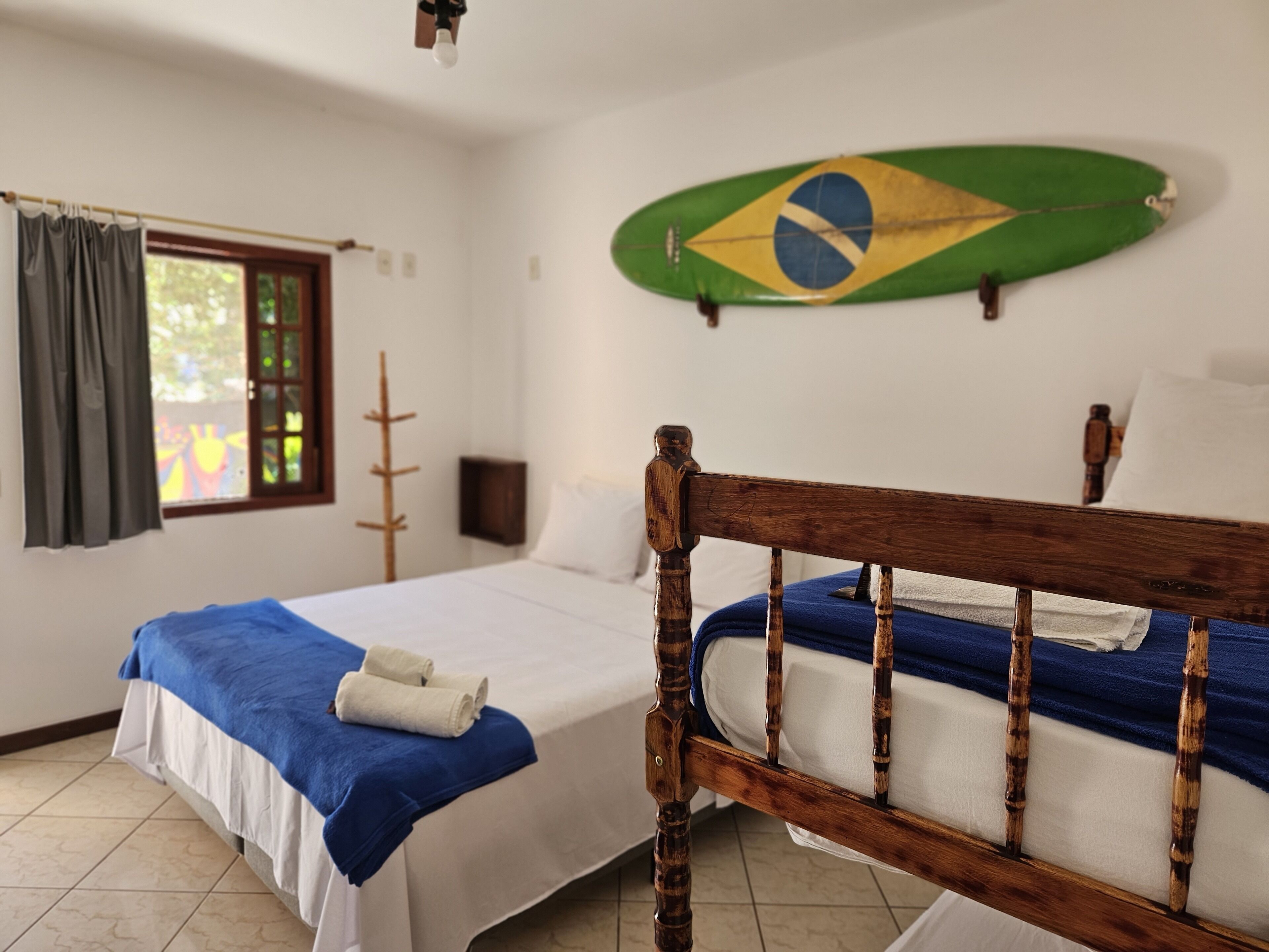 Une chambre avec deux lits, une fenêtre avec des rideaux, une tête de lit en bois et un drapeau brésilien accroché au mur.
