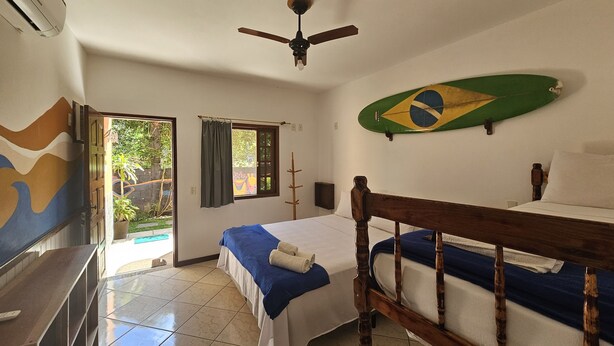 A hotel room with a bed, a ceiling fan, a window with curtains, a wall-mounted surfboard, and a television. - Raio de Sol Pousada & Camping (Angra dos Reis)