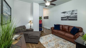 Ein Wohnzimmer mit braunem Ledersofa, grauem Sessel und Hocker, einem gemusterten Teppich und einem Deckenventilator.