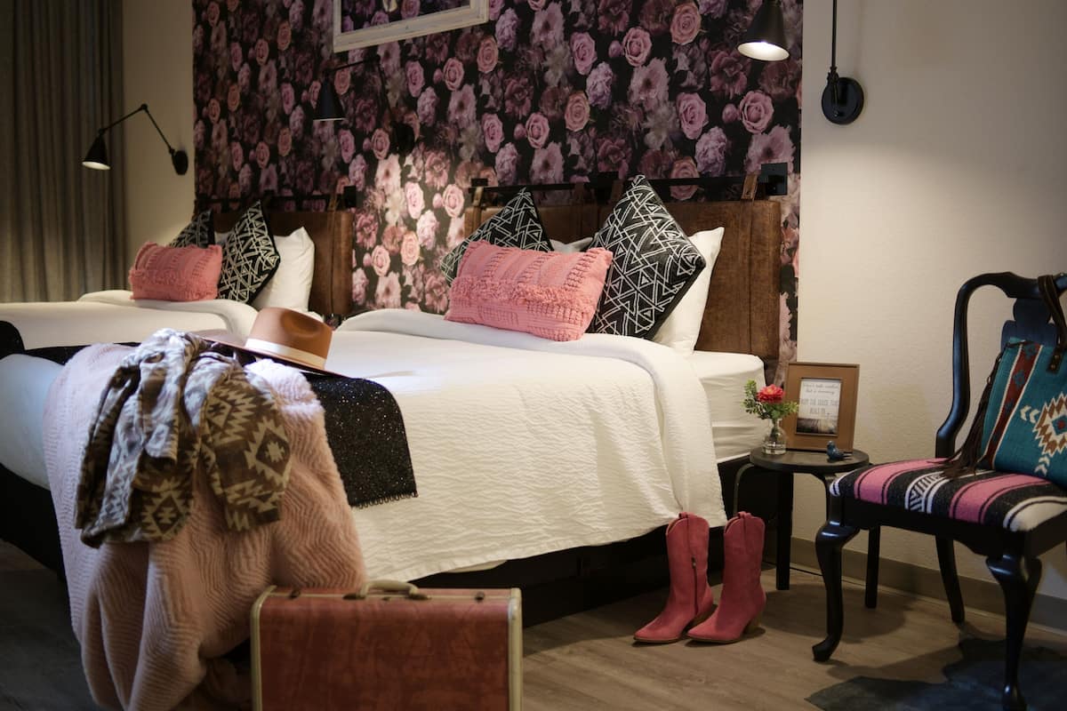 Quarto de hotel com duas camas, papel de parede floral, uma mesinha com um vaso e uma cadeira com almofada estampada.