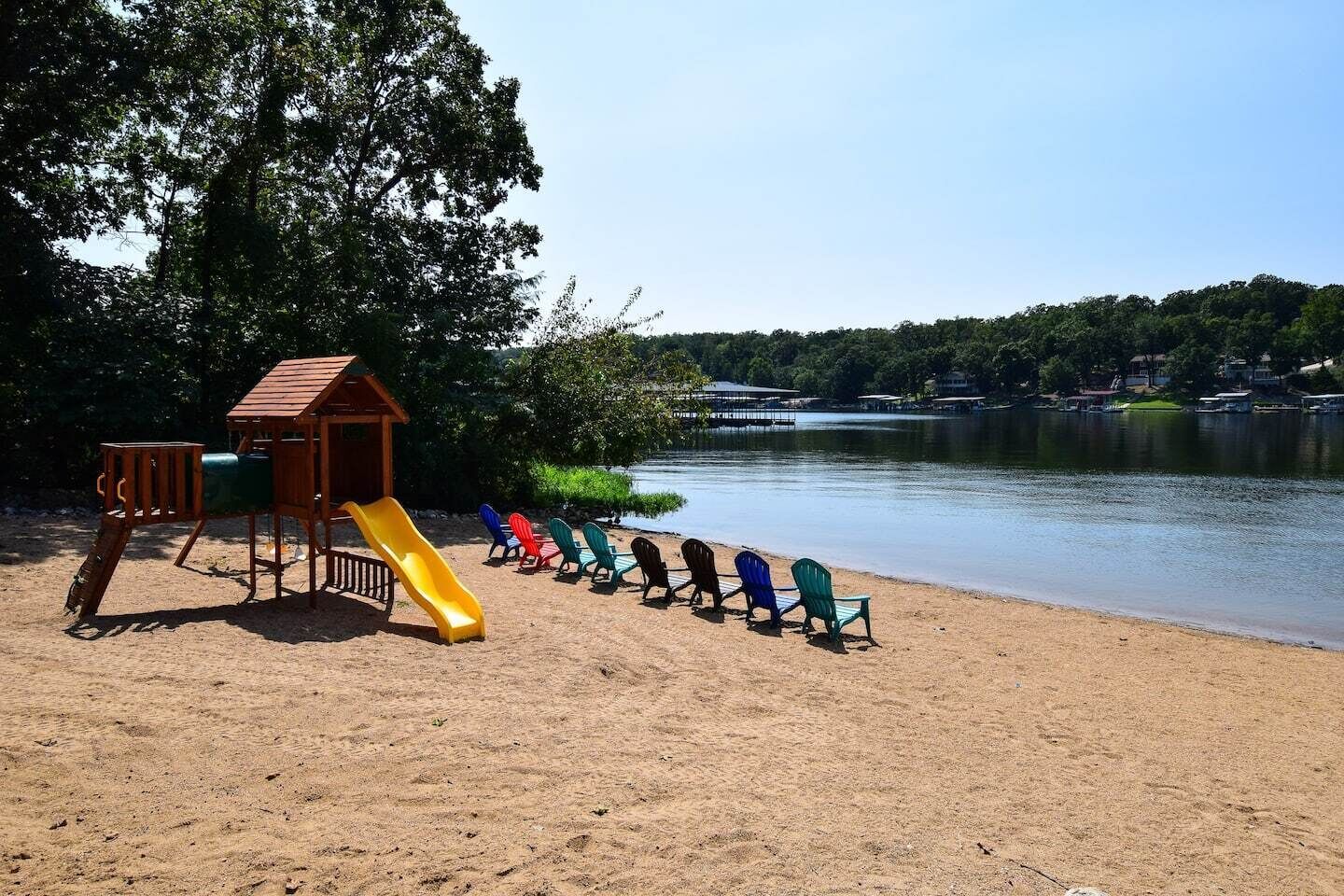Una platja sorrenca amb un parc infantil que inclou un tobogan i una estructura per escalar, i una filera de cadires Adirondack de colors orientades cap a un llac tranquil amb cases al fons.