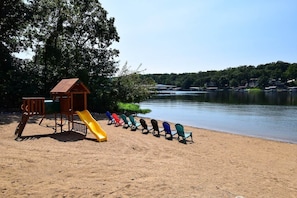Una playa arenosa con un parque infantil que incluye un tobogán y una estructura de escalada, y una fila de sillas Adirondack de colores frente a un lago tranquilo con casas al fondo.