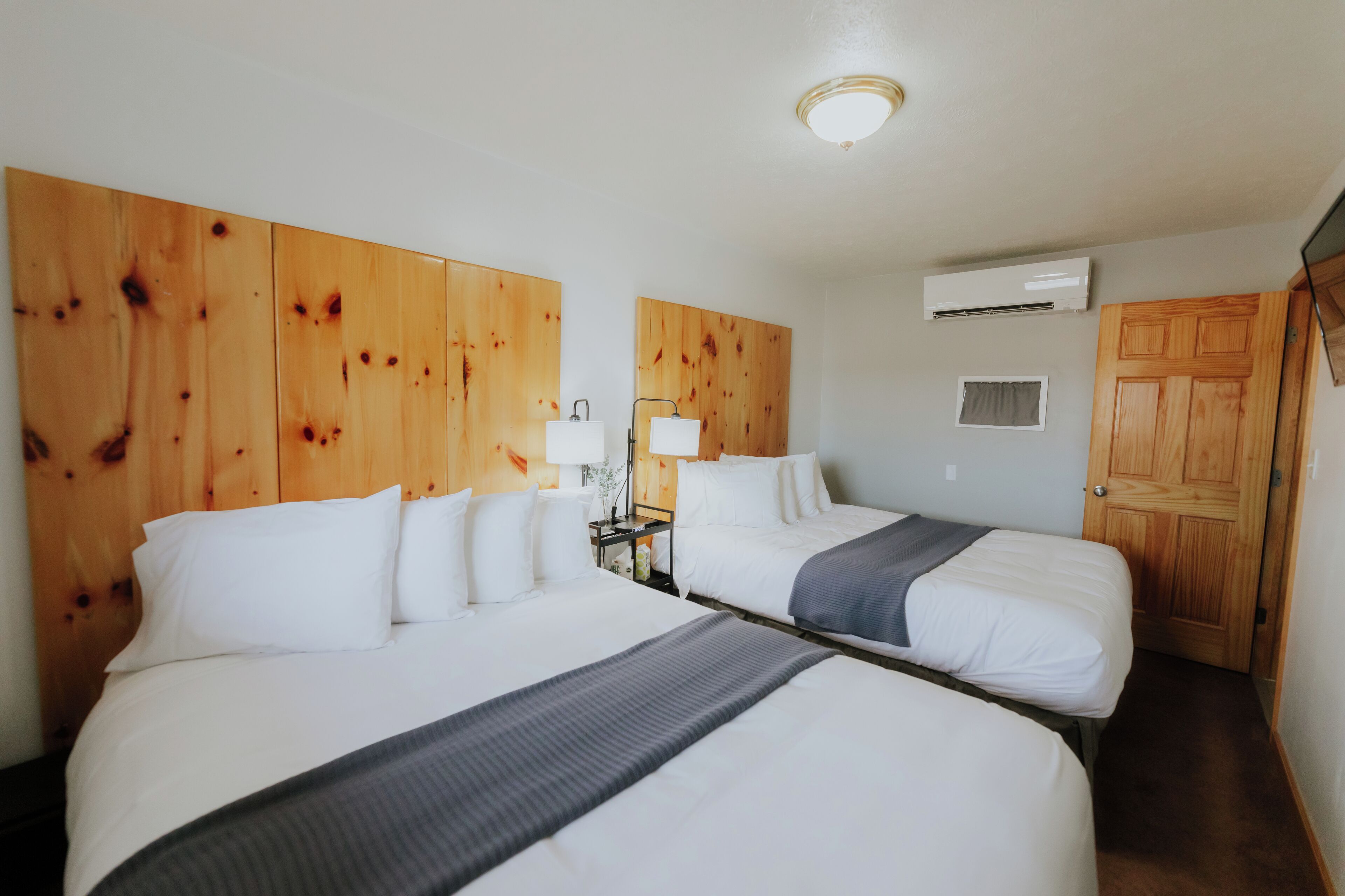 Habitación de hotel con dos camas, cabeceros de madera, ropa de cama blanca, una mesita de noche con lámpara, un equipo de aire acondicionado y una puerta de madera.