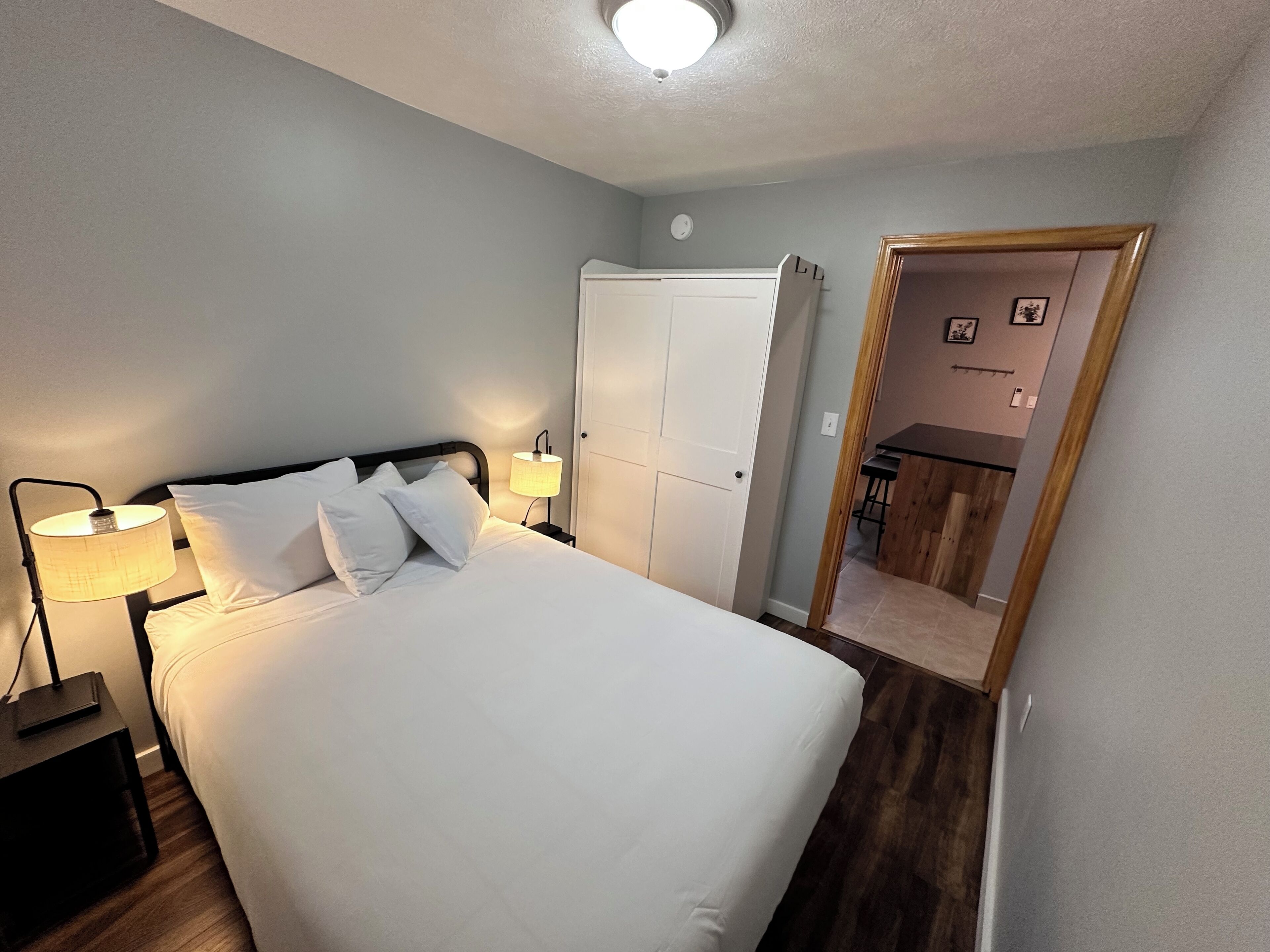 Un lit bien fait, avec du linge de lit blanc et des oreillers, une lampe de chevet, une armoire et un miroir qui reflète un petit coin cuisine.