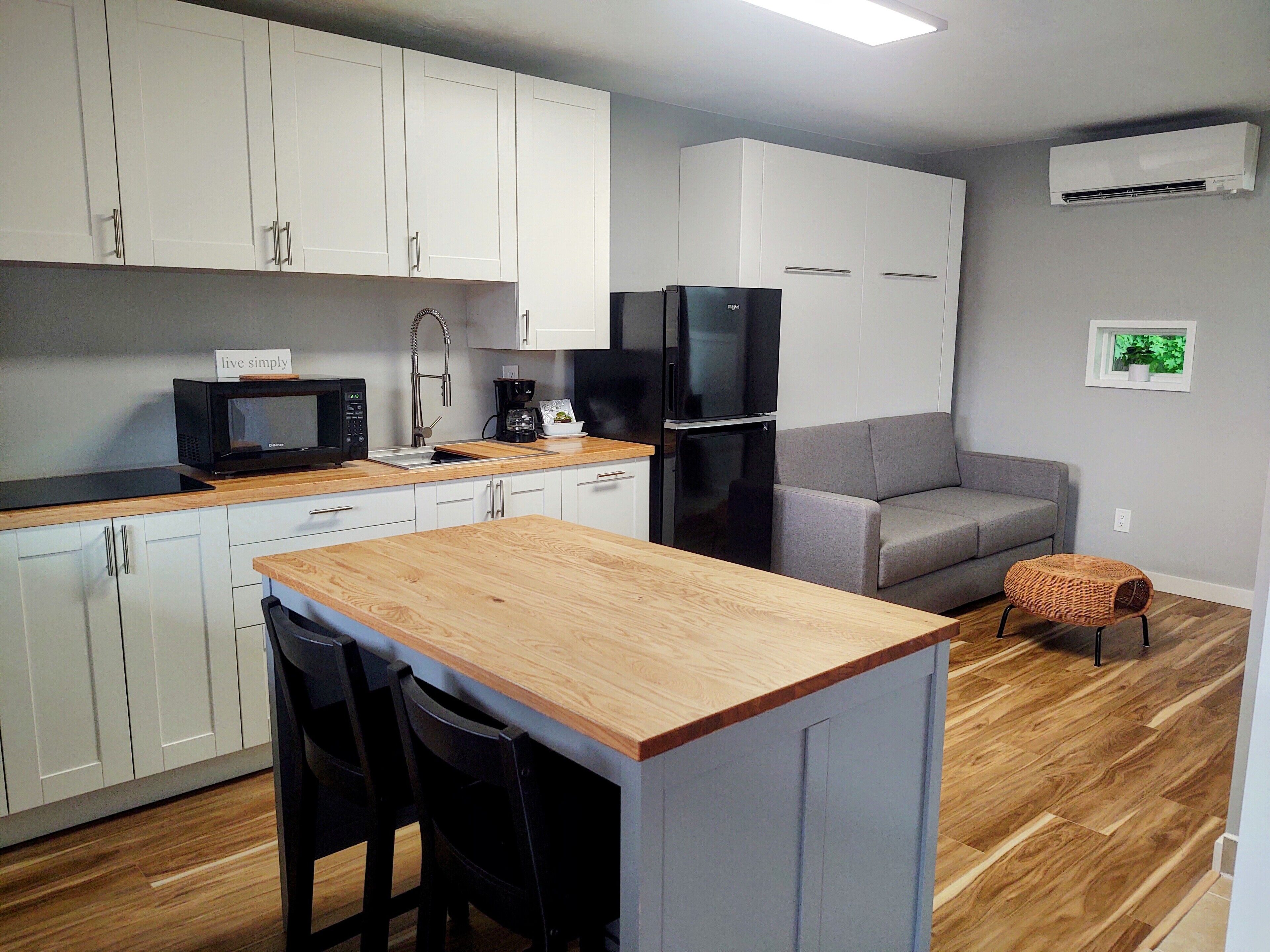 A compact kitchen with white cabinets, a black refrigerator, a microwave, and a coffee maker. There is a wooden island with two chairs and a grey sofa in the living area.