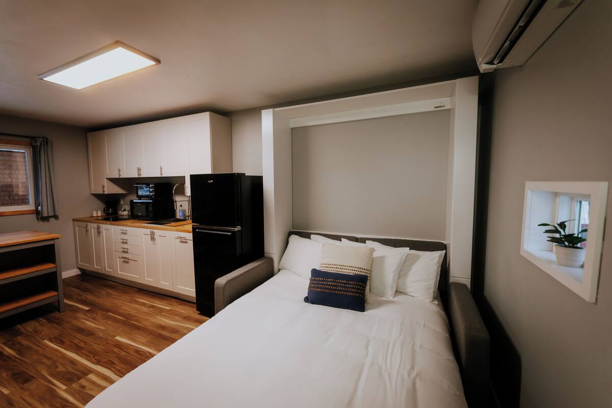 Une chambre d’hôtel compacte, dotée d’une kitchenette, d’un lit avec du linge de lit blanc et d’une petite fenêtre agrémentée d’une plante.