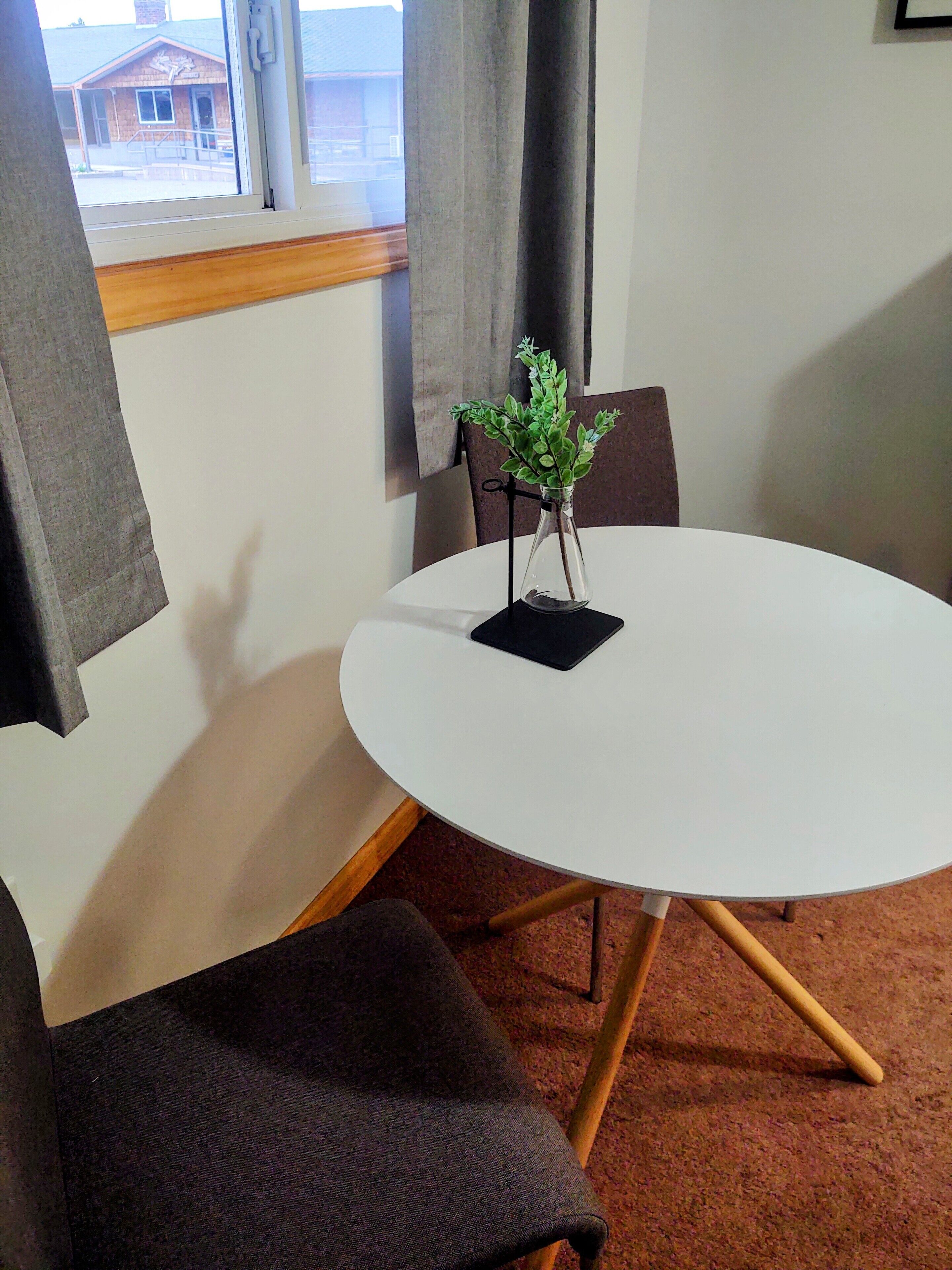 Uma mesa redonda branca com um apoio para copos preto e um pequeno vaso com folhagem verde. Há duas cadeiras marrom-escuro com pernas de madeira. O quarto tem uma janela com cortinas cinzas, e uma vista de um edifício lá fora.