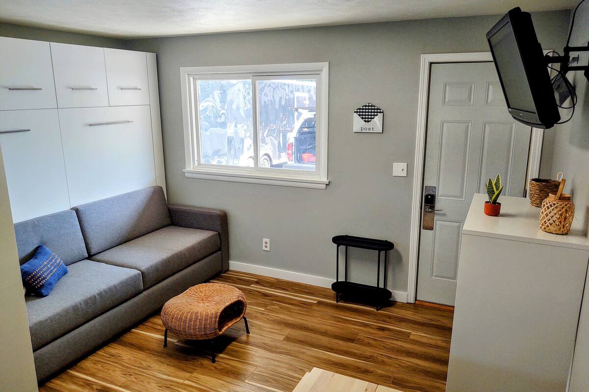 Un espace de vie compact comprenant un canapé gris, un pouf en osier, un petit tabouret noir et un téléviseur fixé au mur.