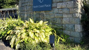 A sign for North Coast Place with a house icon, set against a stone wall with greenery in front. - North Coast Place (Marquette)