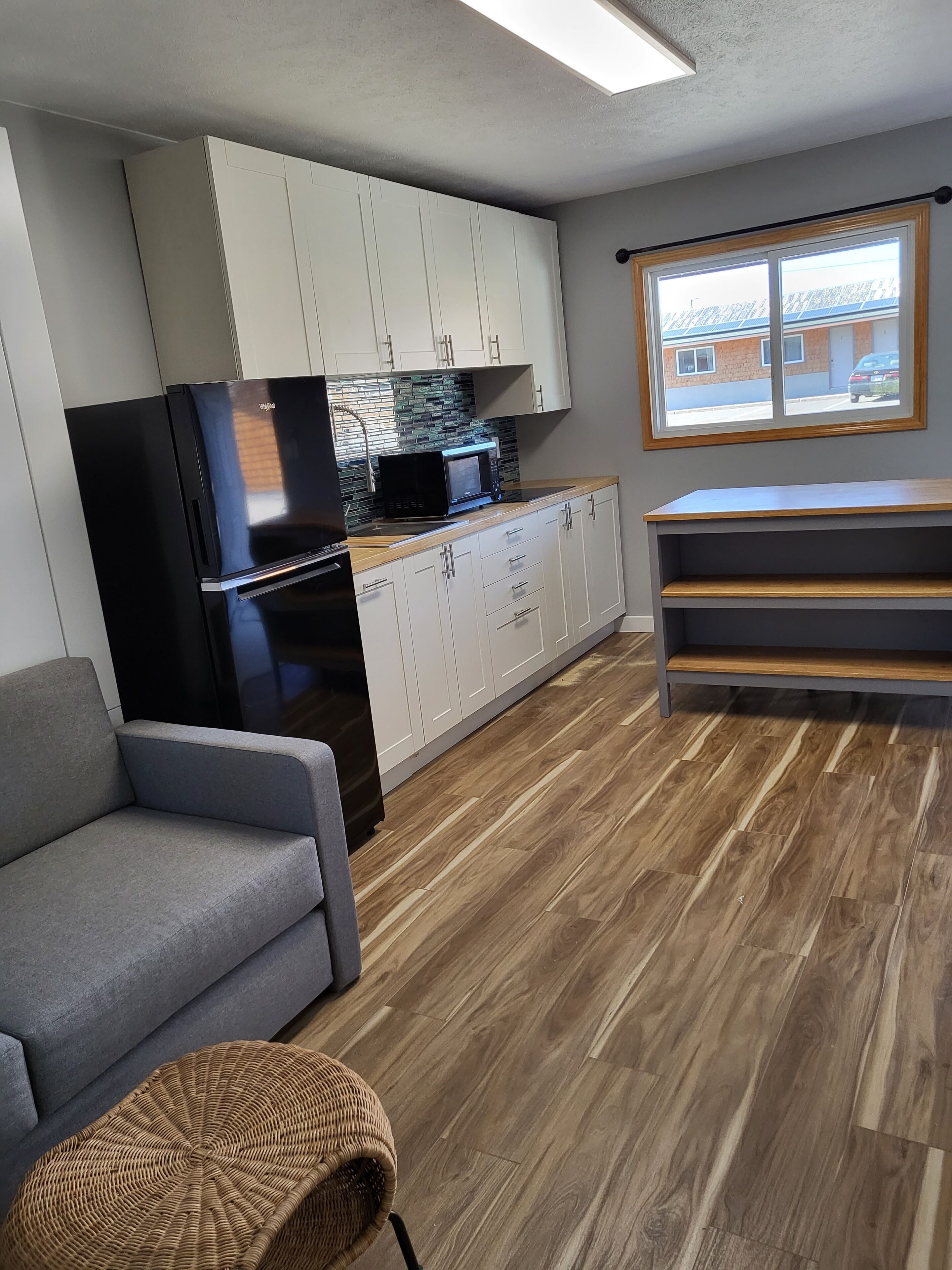 Una cocina moderna con armarios blancos, un refrigerador negro y un microondas. En la sala de estar, hay un sofá gris y un pouf tejido.