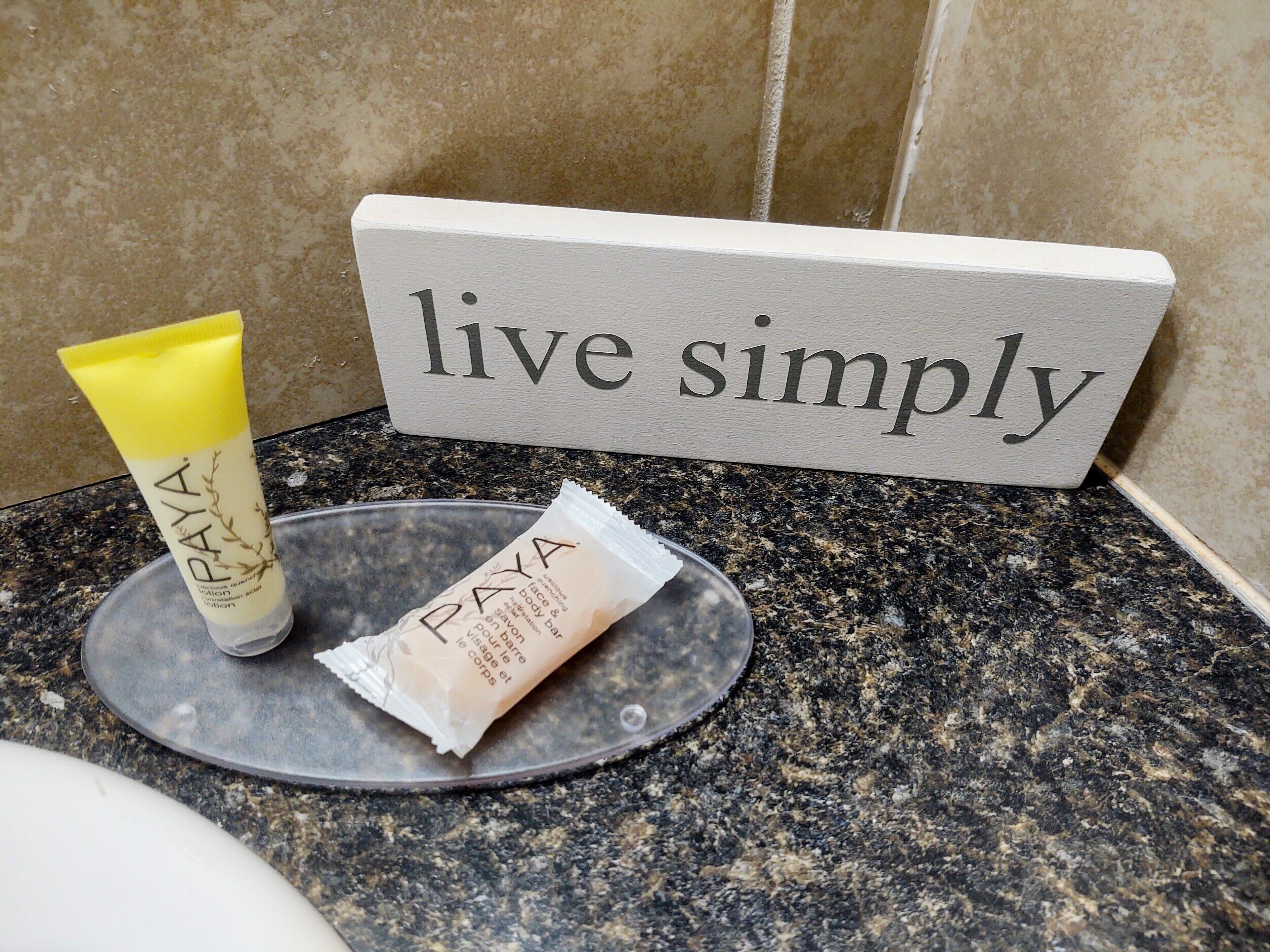 A bathroom counter with a 'live simply' sign, a tube of Paya lotion, and a Paya cream packet.