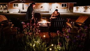 Two people sitting by a fire pit in an outdoor area with wooden chairs and a building in the background. - North Coast Place (Marquette)