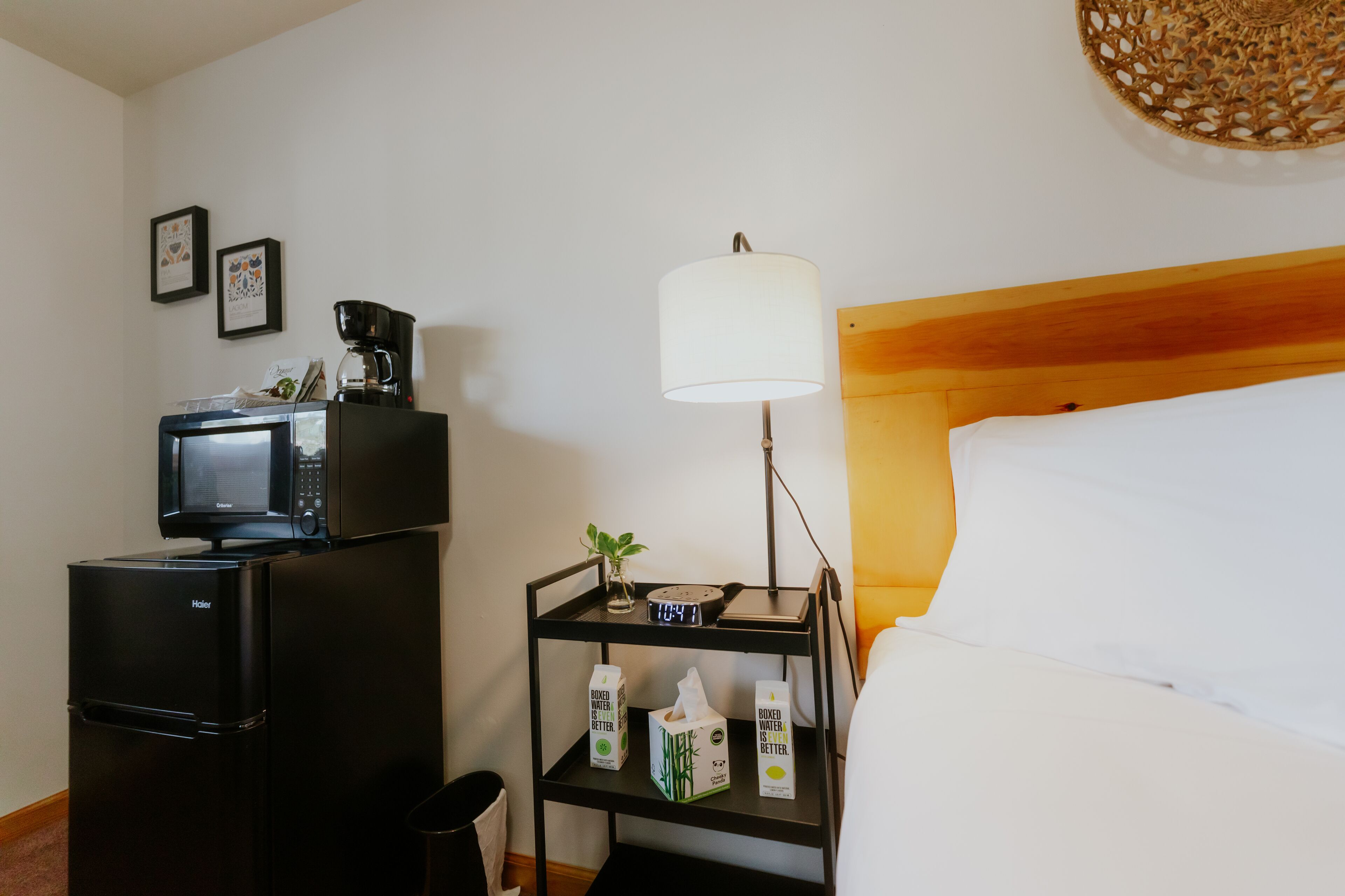 A hotel room with a bed, a bedside table with a lamp, a small shelf with a clock and a plant, a refrigerator, and a microwave.