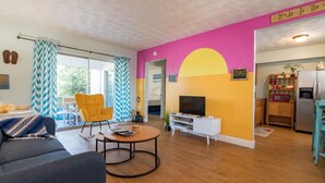Ein Wohnzimmer mit einer Couch, einem gelben Sessel, einem Fernseher und einer Küchenzeile mit einem Kühlschrank.