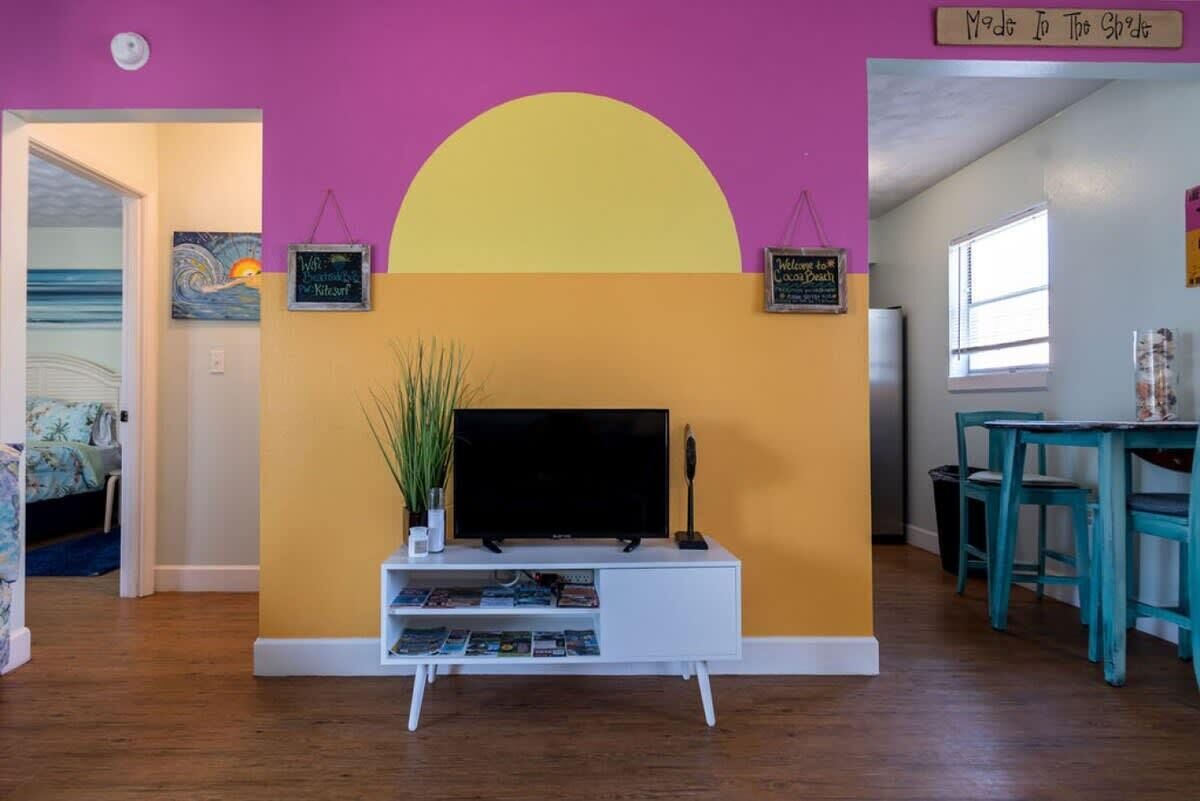 Ein Wohnzimmer mit einem Fernseher auf einem weißen Schrank, einer großen gelben Wand und einer violetten Wand. An der Wand befinden sich zwei Schilder, eines mit der Aufschrift 'Welcome to the Beach' und das andere mit 'Made to the Shore'.