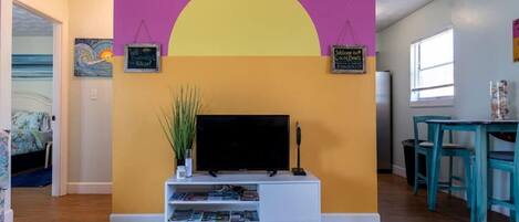 Ein Wohnzimmer mit einem Fernseher auf einem weißen Schrank, einer großen gelben Wand und einer violetten Wand. An der Wand befinden sich zwei Schilder, eines mit der Aufschrift 'Welcome to the Beach' und das andere mit 'Made to the Shore'.