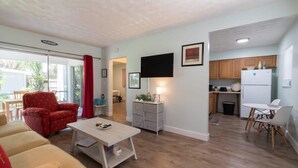 A compact living space with a sofa, a coffee table, a TV, a kitchen area with cabinets and a refrigerator, and a sliding glass door leading to an outdoor area. - 1BR Cocoa Beach Home - 1 Min to Beach! (Cocoa Beach)