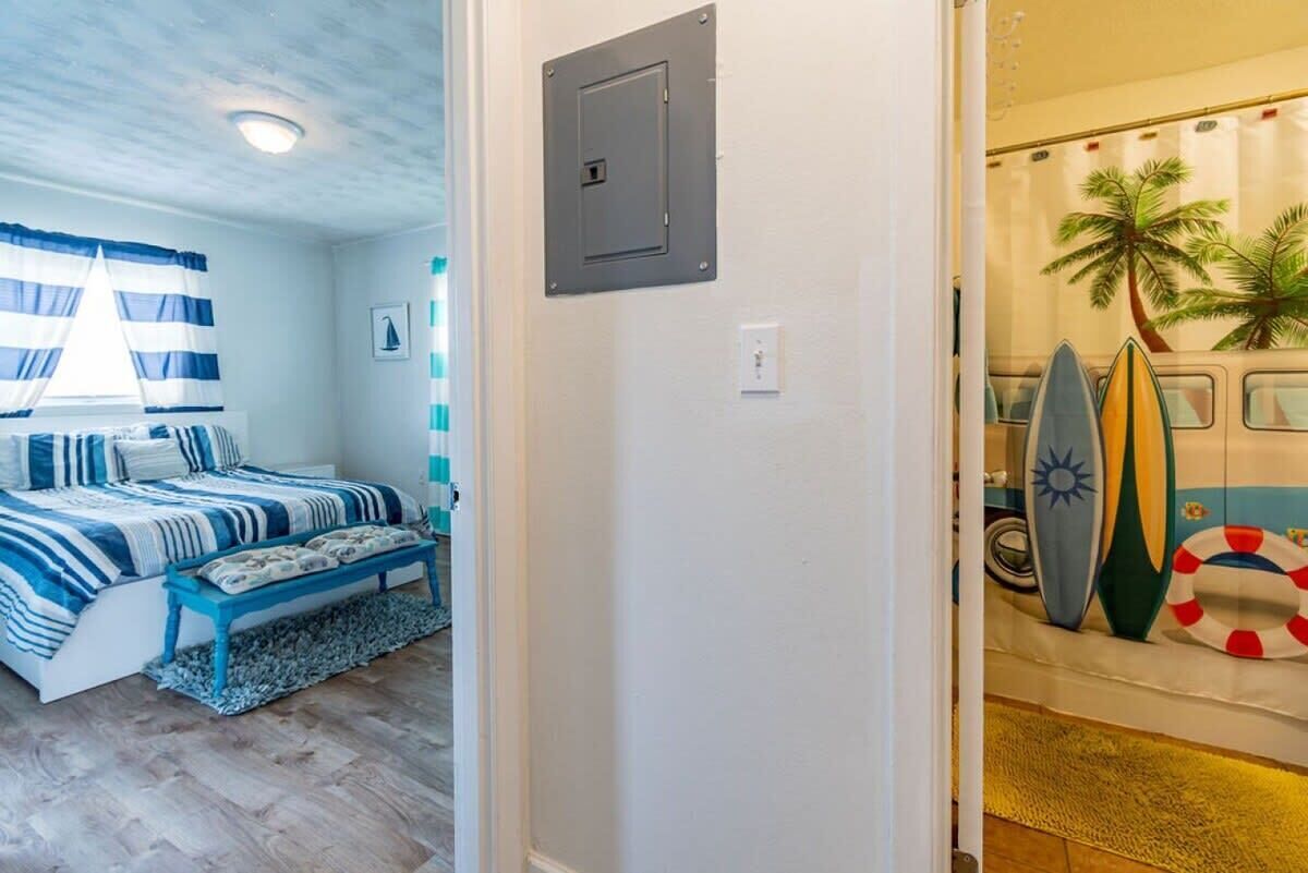 A bedroom with a bed, a bench, a window with curtains, a wall-mounted light, and a door leading to a room with surfboards and a life preserver.
