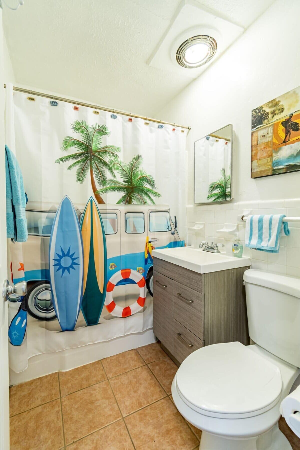 Une salle de bain avec un rideau de douche orné de palmiers et de planches de surf, des toilettes, un lavabo avec un miroir et un porte-serviettes.