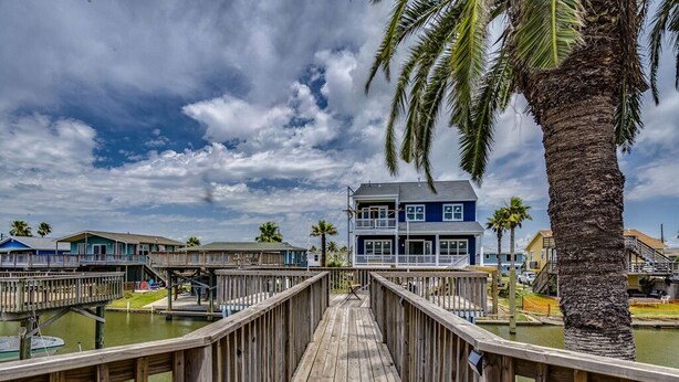 A wooden boardwalk leading to a two-story building with a balcony, surrounded by palm trees and water.