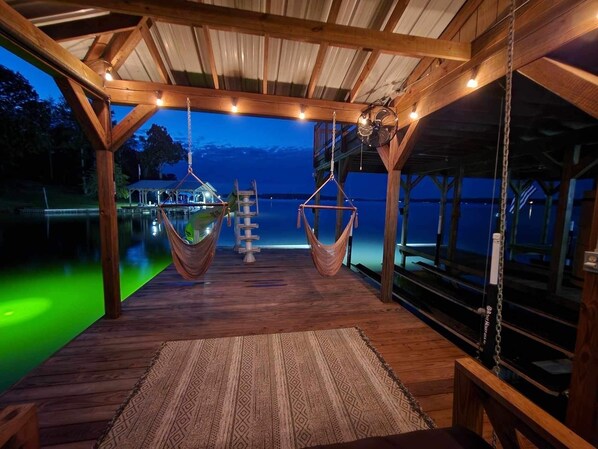 Terraza de madera con dos hamacas colgantes, suelo de madera y vista a un cuerpo de agua al anochecer.