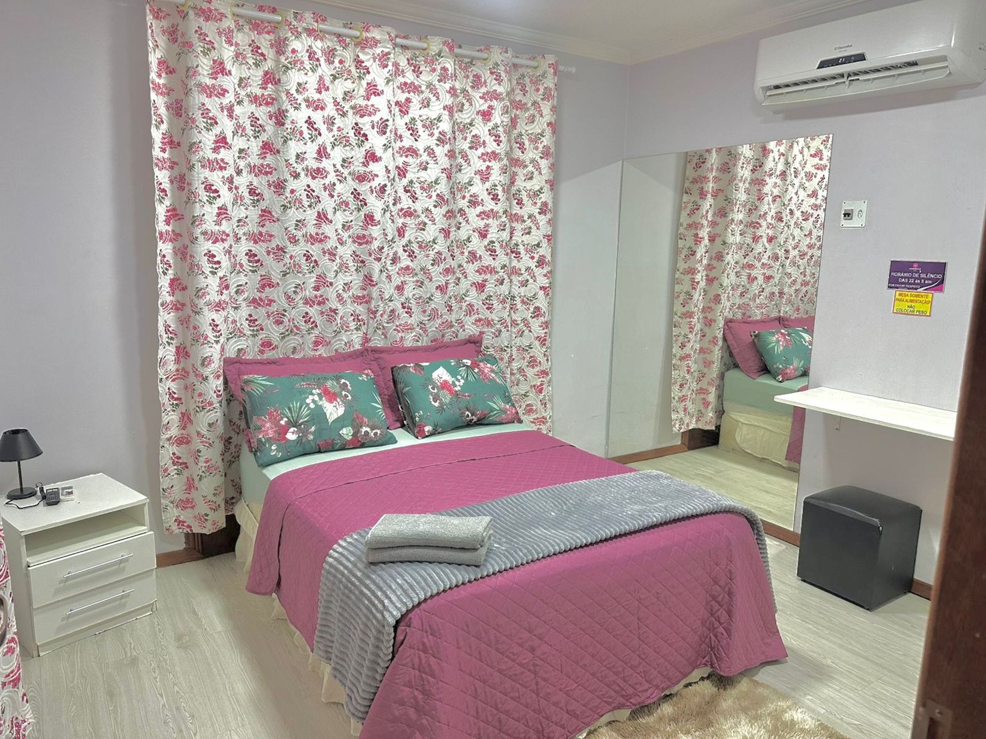 A bedroom with a bed, a bedside table, a mirror, an air conditioning unit, and a small stool.