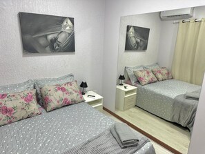Un dormitorio con dos camas, almohadones florales, una mesita de noche con lámpara, un espejo y un aire acondicionado instalado en la pared.