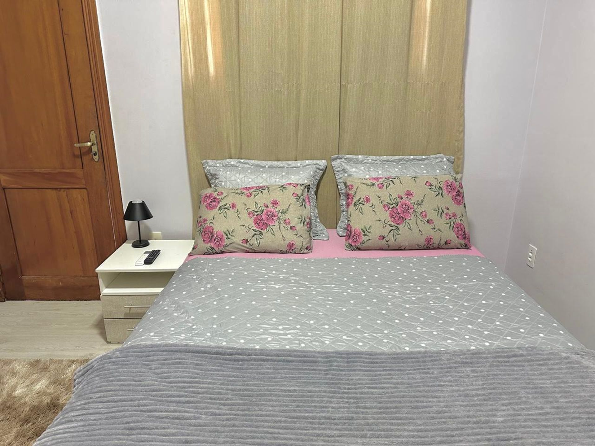 Un dormitorio con cama, dos almohadas, una mesita de noche con lámpara y una puerta de madera.
