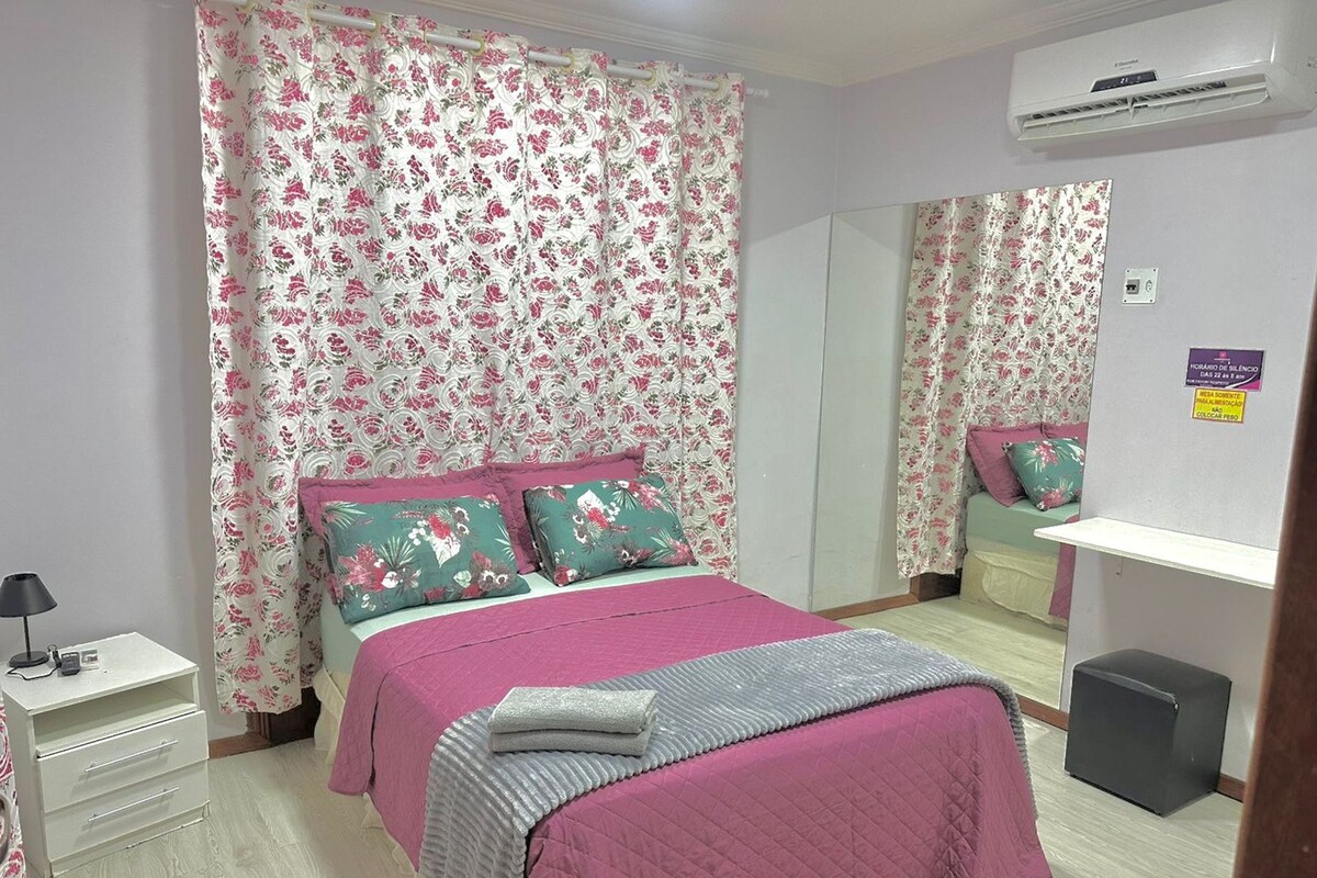 Une chambre à coucher comprenant un lit, une table de chevet, un miroir, un appareil de climatisation et un petit tabouret.